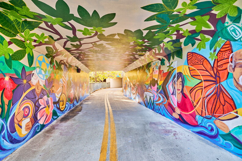 A colorful mural with blues, purples, greens and reds cover the walls and roof of a tunnel. There are flowers, people and a butterfly shown in this shot of the mural.