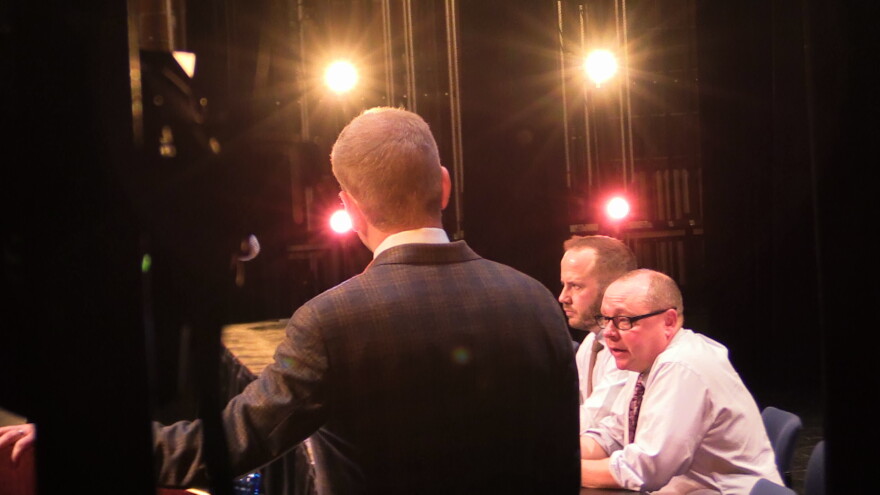 The host of an event looks at two men sitting by a table on stage, with lights shining behind them