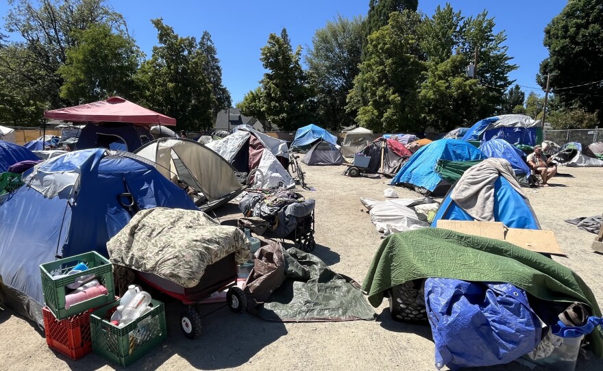 A dirt lot is covered with tents and other belongings, including crates, a wagon and bedding. 