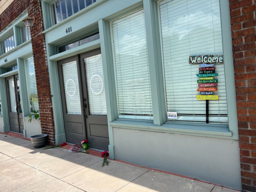A light teal storefront with brick pillars with flowers in front of black doors and closed windows.