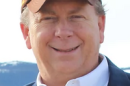 Older man smiling in baseball cap, white button down shirt and black jacket