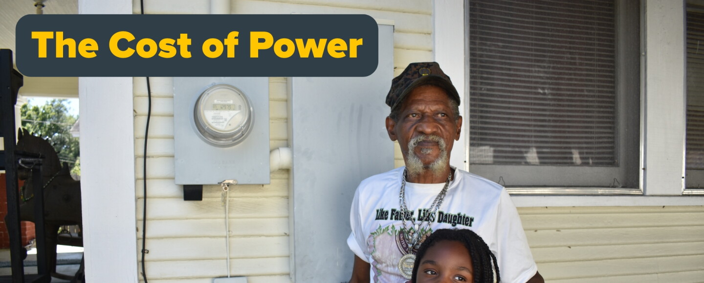 An older Black man with a gray goatee standing on the side of a yellow house, with his left hand on a young girl's shoulder. They're standing in front of a power meter on the house. A grey and yellow text saying "The Cost of Power" to the left 