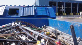 A bin in the foreground is full of metal scraps and cans. There are more blue bins behind it and a large blue pole-barn style building behind it, where trash is dumped.