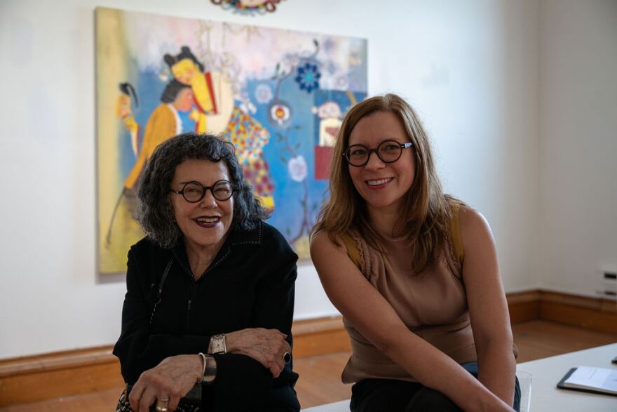 Two women sitting on a white bench with a painting of blue, red, and yellow colors.