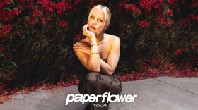 A woman in a sheer dress with a serious expression on her face rests her chin in her hand as she crouches in front of a wall covered in plants.