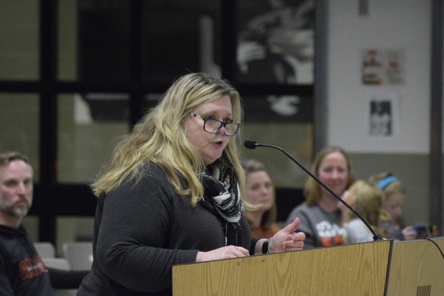 A blonde woman with glasses speaks at a wooden podium.