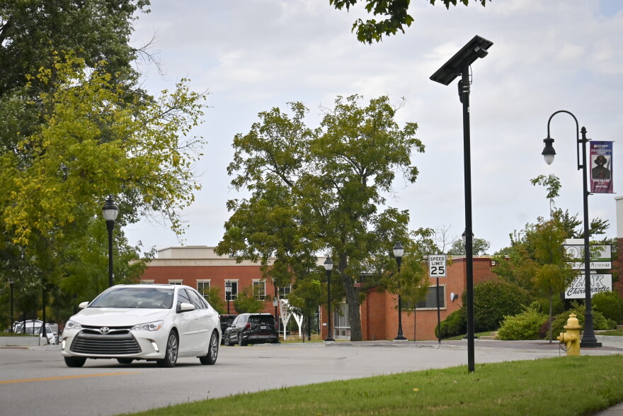 Outdoor photo showing traffic moving on a two-lane street. In background can be seen some brick buildings. In foreground is a black pole with a small solar panel on top and a tiny camera.