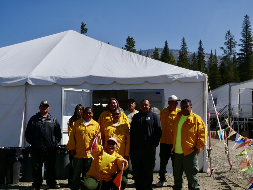 A group of people wearing green pants, yellow button-up shirts and sweatshirts stand together in front of a large white dining tent. A person in front holds up a colorful string of triangular flags and smiles.