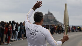 Torchbearer Maxime Gohier waves to the audience as he holds the Olympic torch in front of the Mont-Saint-Michel, Friday, May 31, 2024 in Brittany. (AP Photo/Jeremias Gonzalez)