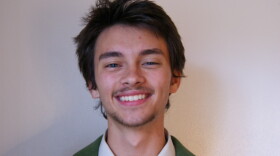 A young man with brown hair and short facial hair wears a green blazer and matching tie. He smiles in front of a white background.