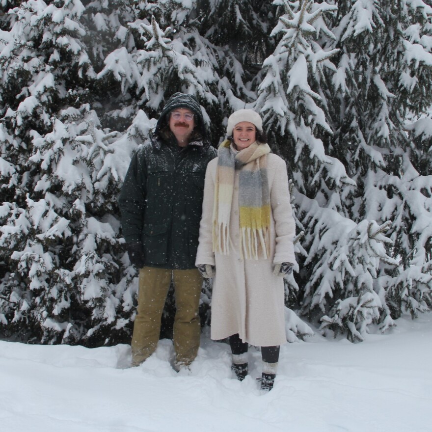 A man and a woman in large winter coats stand in front of a snow covered evergreen tree. 