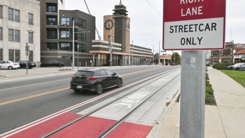 A city street is shown in the background with a single, black car driving one lane. In the lane nearest the camera, a red rectangle can be seen painted on the pavement where streetcar tracks are laid. A red and white sign in the foreground reads "Right Lane, Streetcar Only."