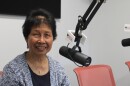 A woman sits behind a hanging microphone at a desk. She smiles at the camera.