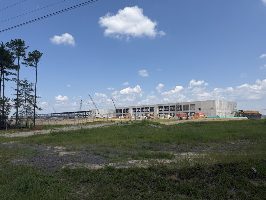 In the center of the photo is a large, concrete building. To the left is the steel skeleton of another giant building. Grass is in the forefront of the picture, and clear skies above the buildings. There are also constructions cranes and other construction equipment. 