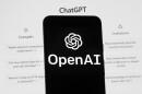 The OpenAI logo is seen on a mobile phone in front of a computer screen which displays the ChatGPT home screen.