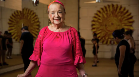 A woman in a hot pink blouse and skirt stands in front of youth dancers wearing black. 