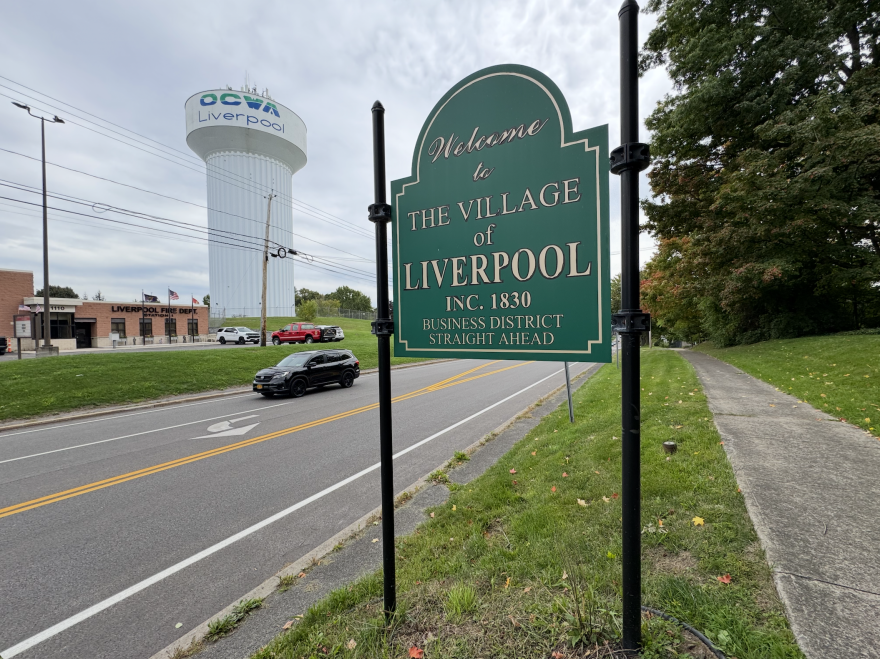 A green sign with gold lettering stands across a two lane road from a Liverpool firestation and water tower. The sign reads, "Welcome to The Village of Liverpool Inc. 1830 Business District Straight Ahead."