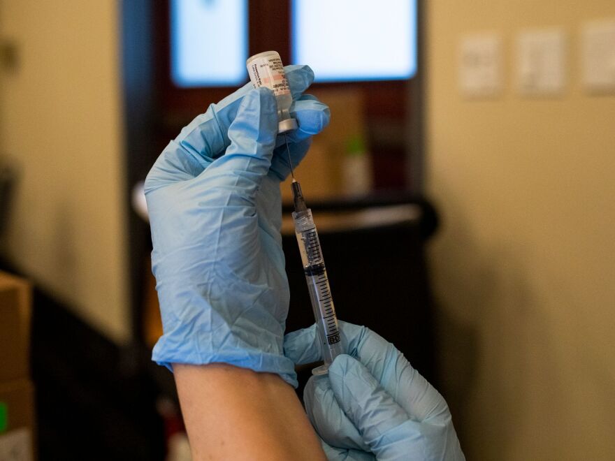 Two hands wearing blue gloves hold up a vial of vaccine with a syringe injected.