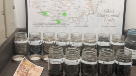 14 jars filled with dirt sit on a shelf in front of a map of the Underground Railroad stops in Ohio. Each jar is labeled with a man's name and a city name. 