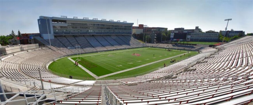 Empty stands surround a green football field. In the distance, the press box casts a shadow over the stands.