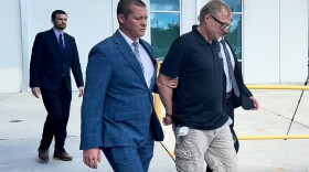 A man in a blue pin striped suit leads a man in handcuffs who is wearing a black t-shirt and cargo shorts off to the right. 
