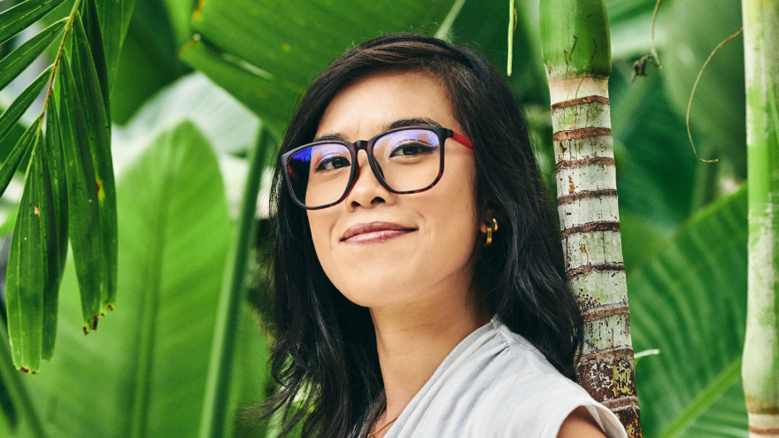 A headshot of Stephanie Foo, wearing glasses with the large green leaves of a growing plant behind her