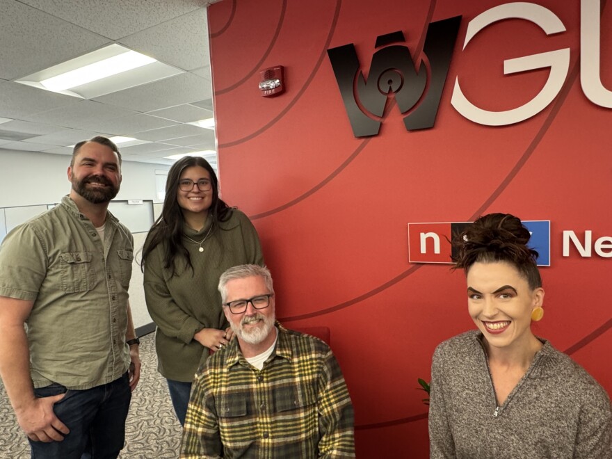 Four people pose and smile in an office near a red wall with the "WGL" logo and a news sign. Three people stand while one man sits, all casually dressed, in a well-lit workspace.