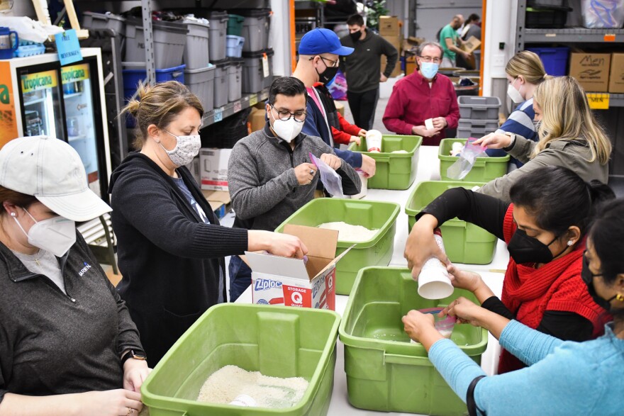 Room full of masked people use cups to pull rice from green bins and fill plastic bags.