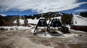 A ski lift surrounded by dirt and some snow further up the mountain on a mostly sunny day.