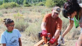 Three people wearing safety glasses and gloves assemble wooden boards outdoors, with one person using a cordless drill.