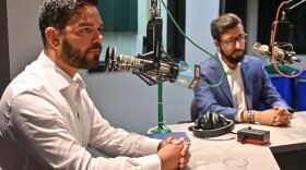 Two men sitting inside a radio studio talk at microphones.
