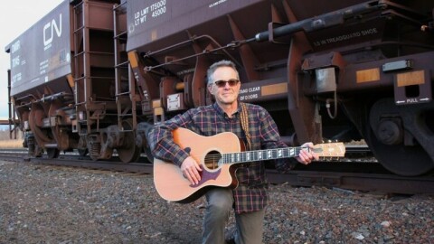 A man with a guitar kneels in the gravel alongside freight train cars