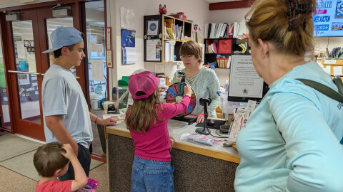 Delta Community Library Director Tiki Levinson, behind counter, helps another family get rewards for reading. Ten-year-old Reilly Testa takes a turn at the prize wheel for reading several books as part of the Delta Community's Library's Summer Reading Program. Her brothers, Mikey, 3, and Peter, 12, wait for their turn as their grandma, Kat Connor, looks on.