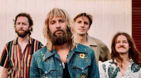 A bearded man in a blue embroidered shirt stands in front of three other band members, all posing outdoors for a group photo.