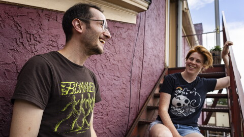 Two people who look to be in their 30s laugh while standing and sitting on the stairs outside a burgundy building.