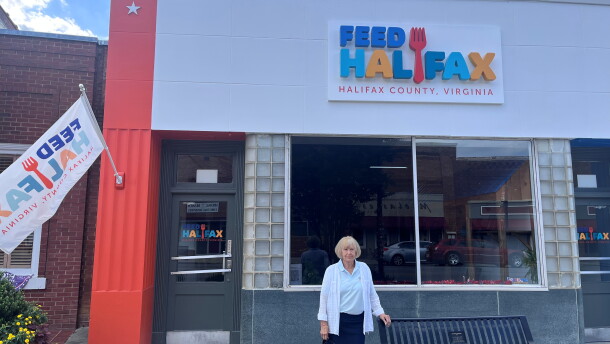 A woman wearing a blue skirt and white cardigan stands in front of a building painted in bright orange on the side and the words "Feed Halifax" on the face of the building. 