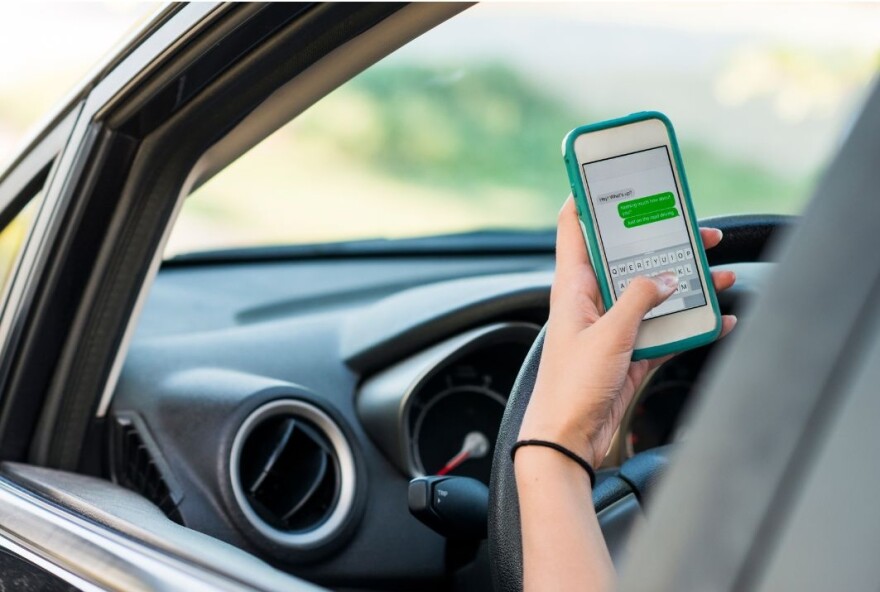 On driver's side of car with steering wheel in view, the hand of the driver is seen holding a smartphone and texting