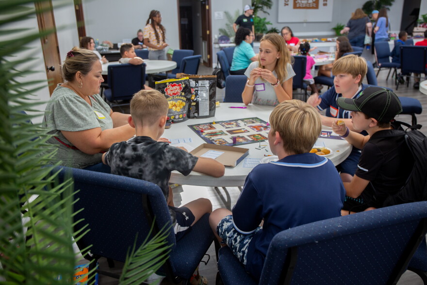 A group of five kids is sitting at a circular folding table with an adult eating chips and popcorn. They're playing the classic board game Clue.