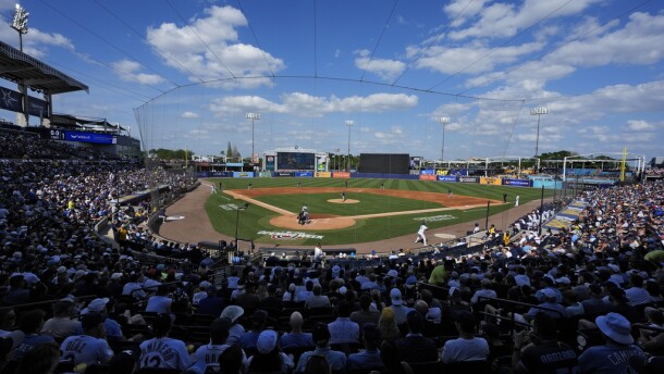 Wide angle image shows a baseball stadium with the crowd filling the seats surrounding the field.  The sky is blue over the outfield with several puffy white clouds. 