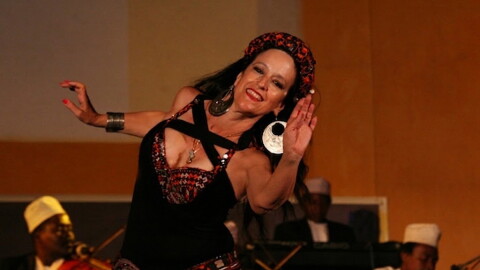 A woman in a black dress with red patterned trim dances in front of a small group of musicians