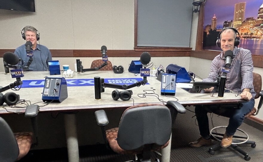 Two smiling men wearing headphones sit at a table in a radio talk studio: the man at left has short grey hair and is wearing a blue button-down shirt; the man at right has short dark hair and is wearing a purple and white checked button-down shirt, jeans, and brown shoes