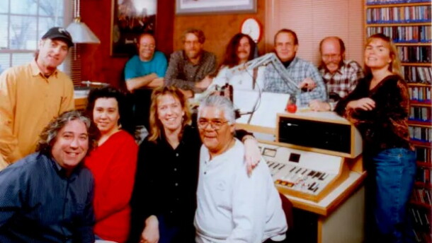 A group of 11 smiling people face the camera, they are seated and standing in a radio control room with a control board and a shelf of CDs in the back.
