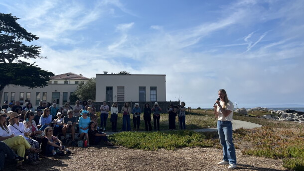 A woman stands holding a microphone with seated and standing audience members looking at her on the edge of the coast. 