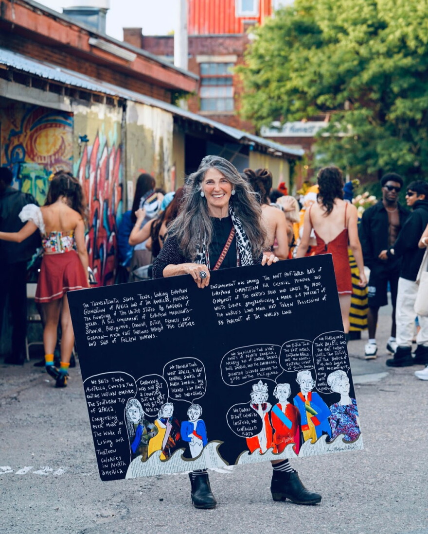 A woman with long grey and brown hair stands in front of a crowd outside holding an oversized book page filled with illustrations of colonizers