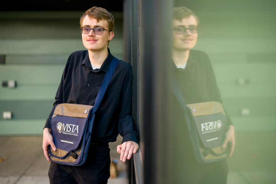 Austin Draper stands next to a reflective wall wearing a blue and brown bag with the VISTA logo.