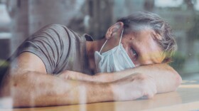 Man in a white protective mask, looking outside through a window,during recovery from COVID illness in home.