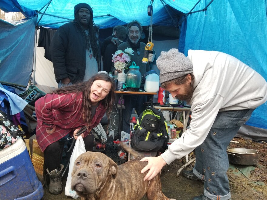 Four smiling people and a dog are in a tent made of blue tarps