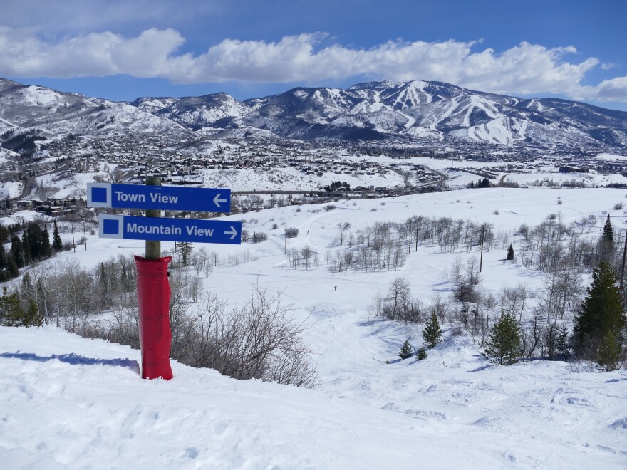On a mountain in Steamboat Springs, two blue signs sit on top of a pole wrapped with red padding. The blue sign on top reads "Town View" with an arrow pointing to the left. The blue sign on the bottom reads "Mountain View" with an arrow pointing to the right. Snow-covered mountains and a blue sky are in the background