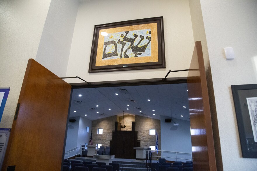 Two large wooden doors are open to reveal the sanctuary of a synagogue, a simple but elegant white-painted room. A framed picture with "Shalom" written in Hebrew hangs above the door.