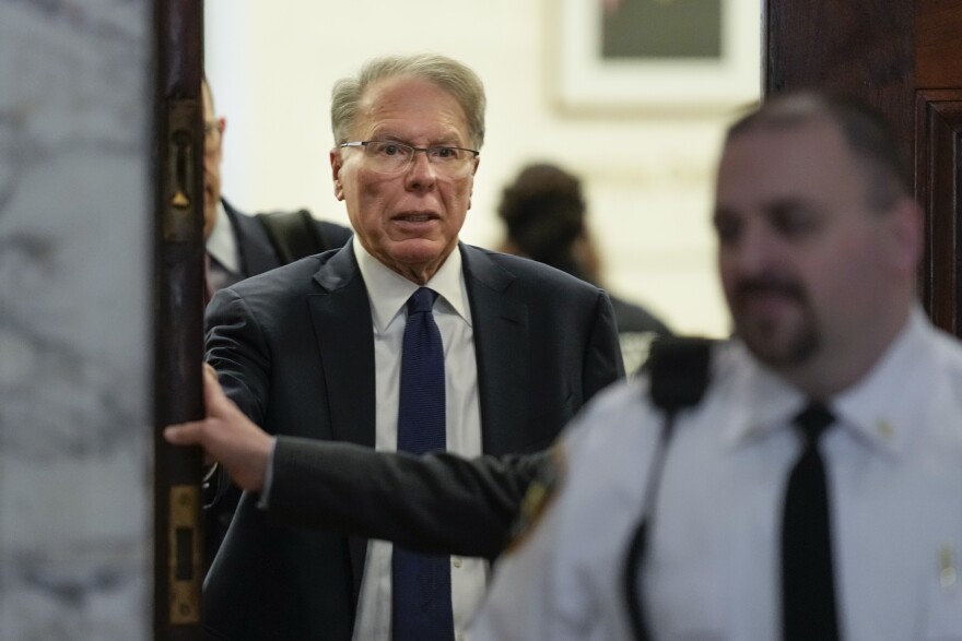 A man in glasses, dressed in a dark suit, stands in a doorway behind a security officer.